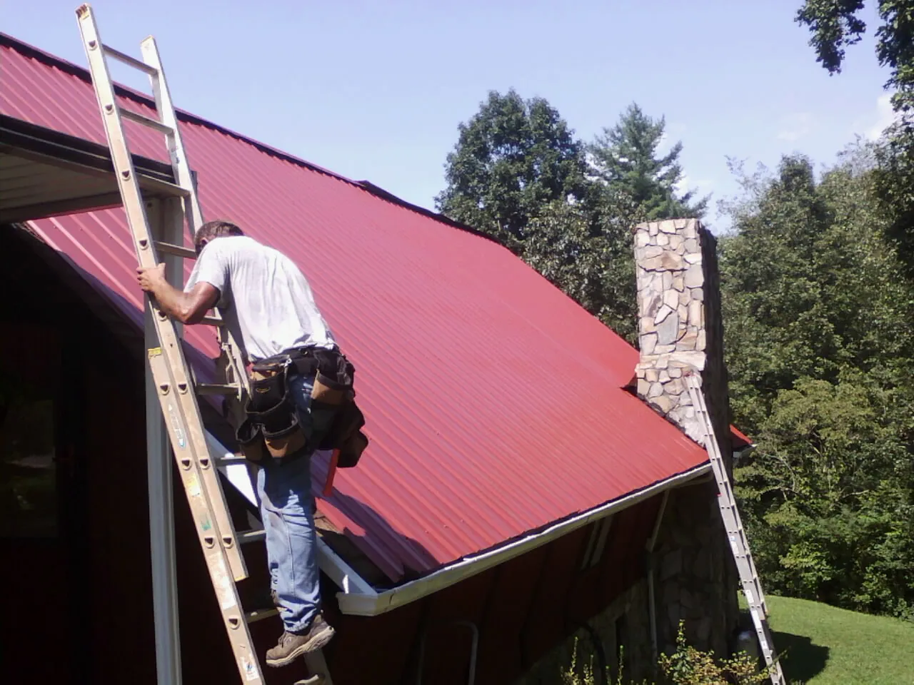 Roofer on a ladder beside a red metal roof and stone chimney — Soffit & Fascia Repair by Roofing Brothers in Santo Domingo Pueblo
